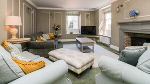Een woonkamer met een bank, fauteuil en voetenbankje, een televisie, een open haard en een raam met gordijnen.