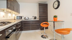 A modern kitchen with dark wood cabinets, a gas stove, a microwave, and a toaster. There is a round orange chair and a small orange table with a potted plant. - 60 Galley Hill View (Bexhill-on-Sea)