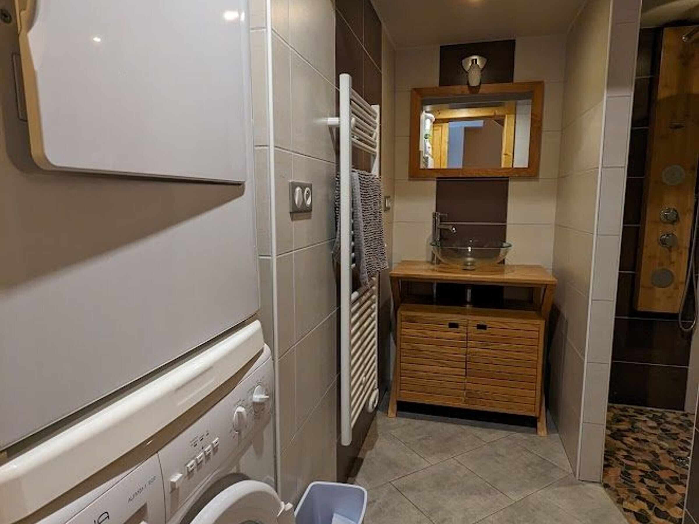 A compact bathroom with a washing machine, a wooden vanity, a mirror, and a shower area.