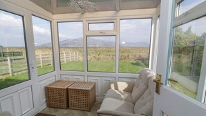 A conservatory with a view of a grassy field and mountains, wooden crates, and a cushioned bench. - Hafod-y-gors (Fairbourne)