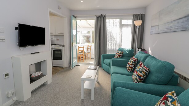 A modern living room with a teal sofa set, a flat-screen TV mounted on the wall, a small white coffee table, and a kitchen area visible through an open door. - Hafod-y-gors (Fairbourne)