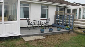 A mobile home with a patio area featuring a table and chairs, potted plants, and a wooden fence. - Hafod-y-gors (Fairbourne)
