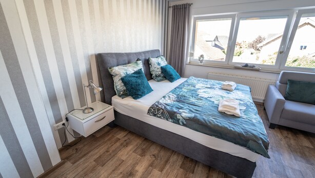A bedroom with a large bed, a bedside table, a chair, and a window with a view of the outside. - Ferienwohnung Anke - Appartement 5c (Heinsberg)