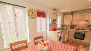 A compact kitchen with a dining table set for four, a washing machine, and a red clock on the wall. - White Goose Cottage (Whitby)