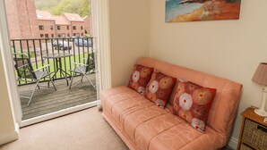 A cozy room with a sofa, a painting of a beach, and a balcony with chairs and a table. - White Goose Cottage (Whitby)