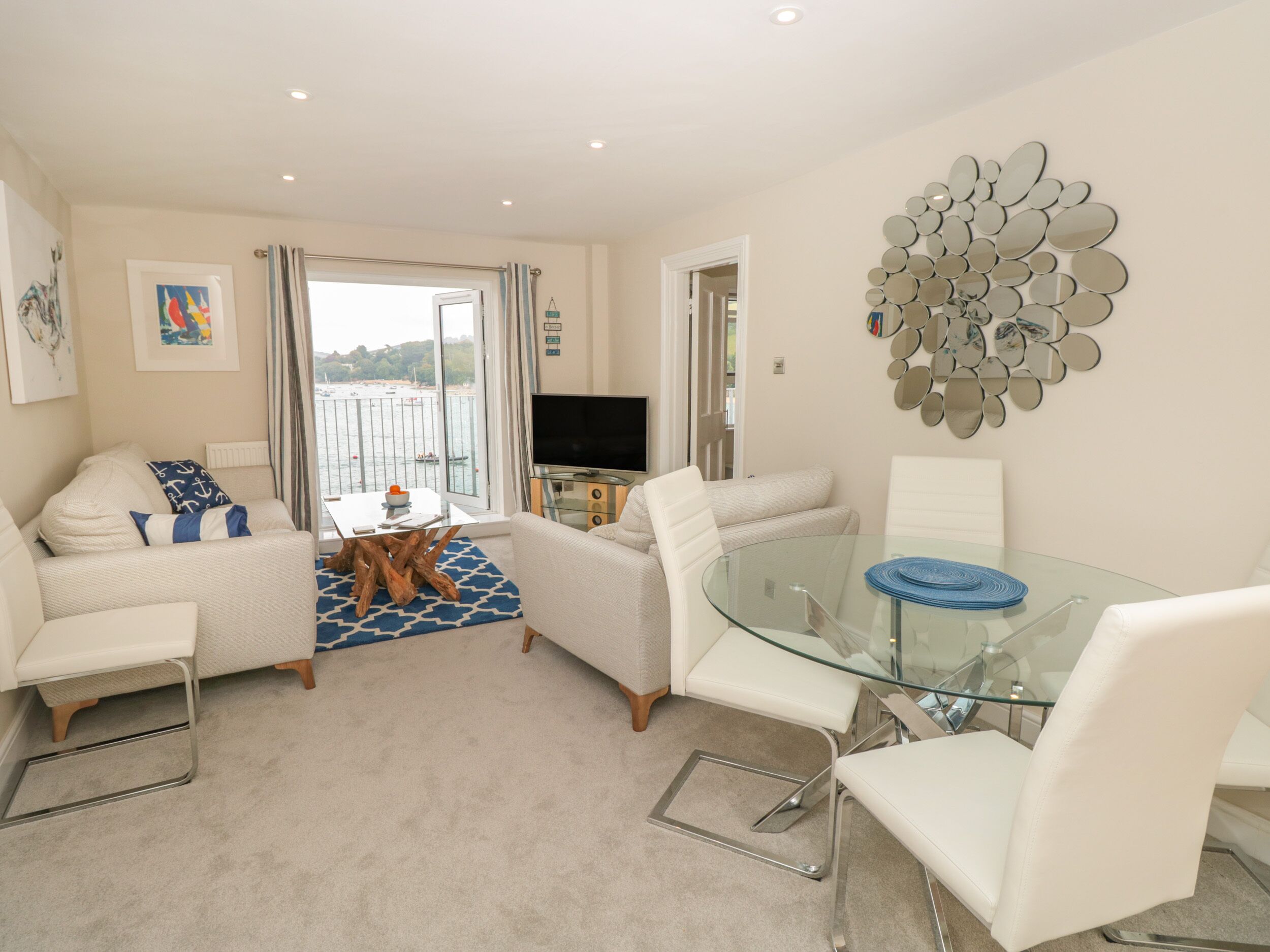 A modern living room with a glass dining table, white sofas, and a view of the sea through large windows.