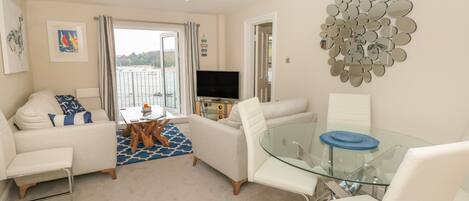A modern living room with a glass dining table, white sofas, and a view of the sea through large windows.