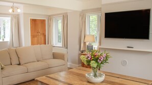 A living room with a beige sofa, a wooden coffee table with a vase of flowers, a television on the wall, and a window with curtains. - The Big Dingle (Church Stretton)