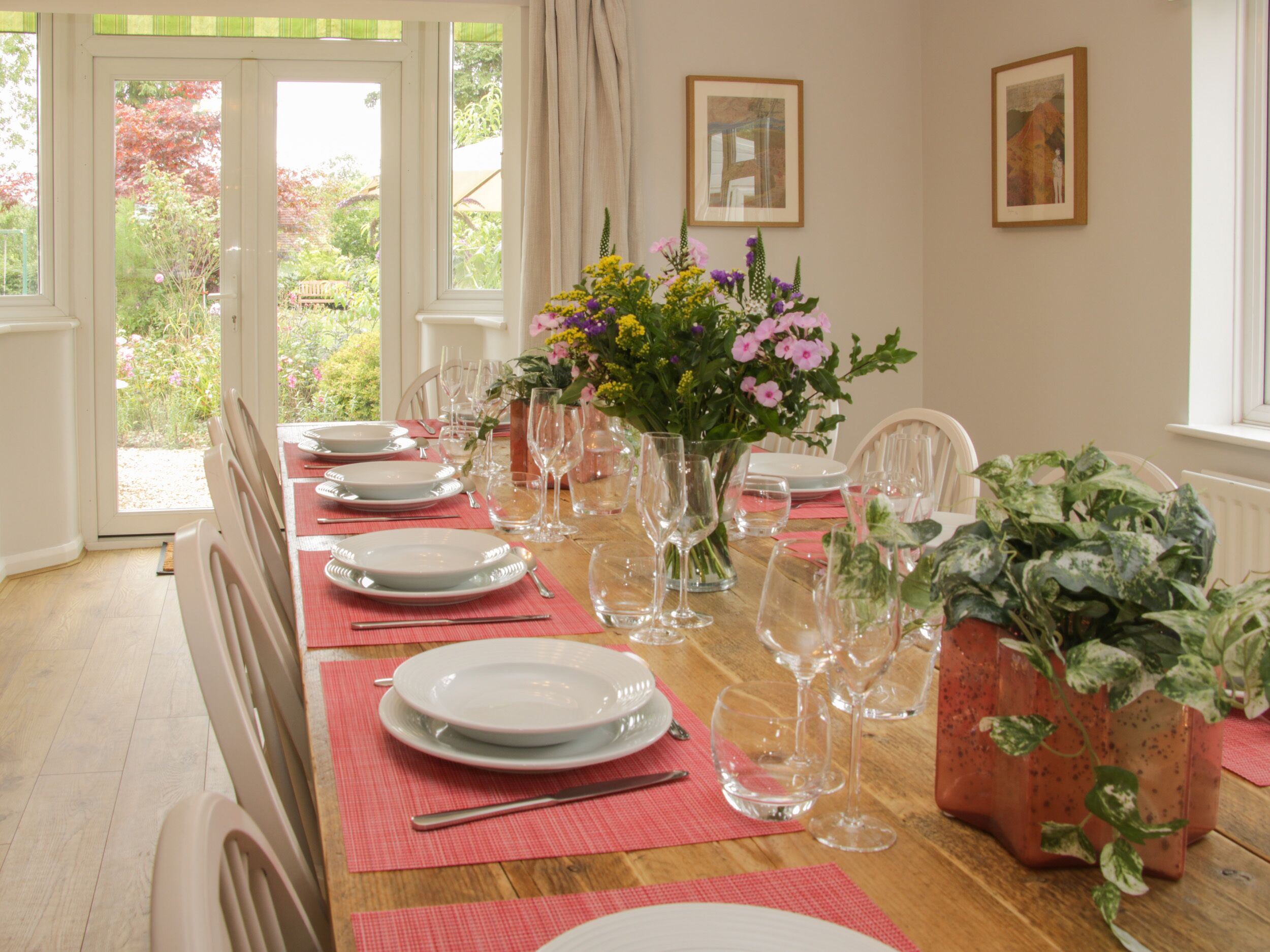 Une table à manger dressée pour six personnes, avec assiettes, couverts et verres à vin, entourée de plantes en pot et de tableaux encadrés accrochés aux murs.