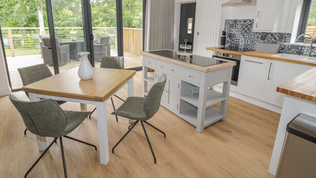 A modern kitchen with a wooden table and chairs, a built-in stove, and a large window overlooking a deck. - The Barn (Redditch)