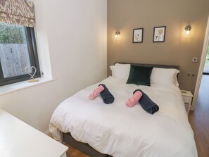 A neatly made bed with white bedding and two rolled towels, a bedside table, a window with a curtain, and wall-mounted lights. - The Leat (Redditch)