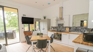 A modern kitchen with white cabinets, a wooden countertop, and a built-in oven. There is a dining area with a table and chairs, and a TV mounted on the wall. - The Leat (Redditch)