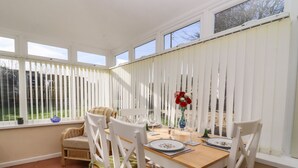 Un coin repas avec une table en bois dressée pour quatre personnes, des chaises blanches avec des coussins verts, un vase rempli de fleurs rouges et, à travers des stores blancs, une vue sur l’extérieur.