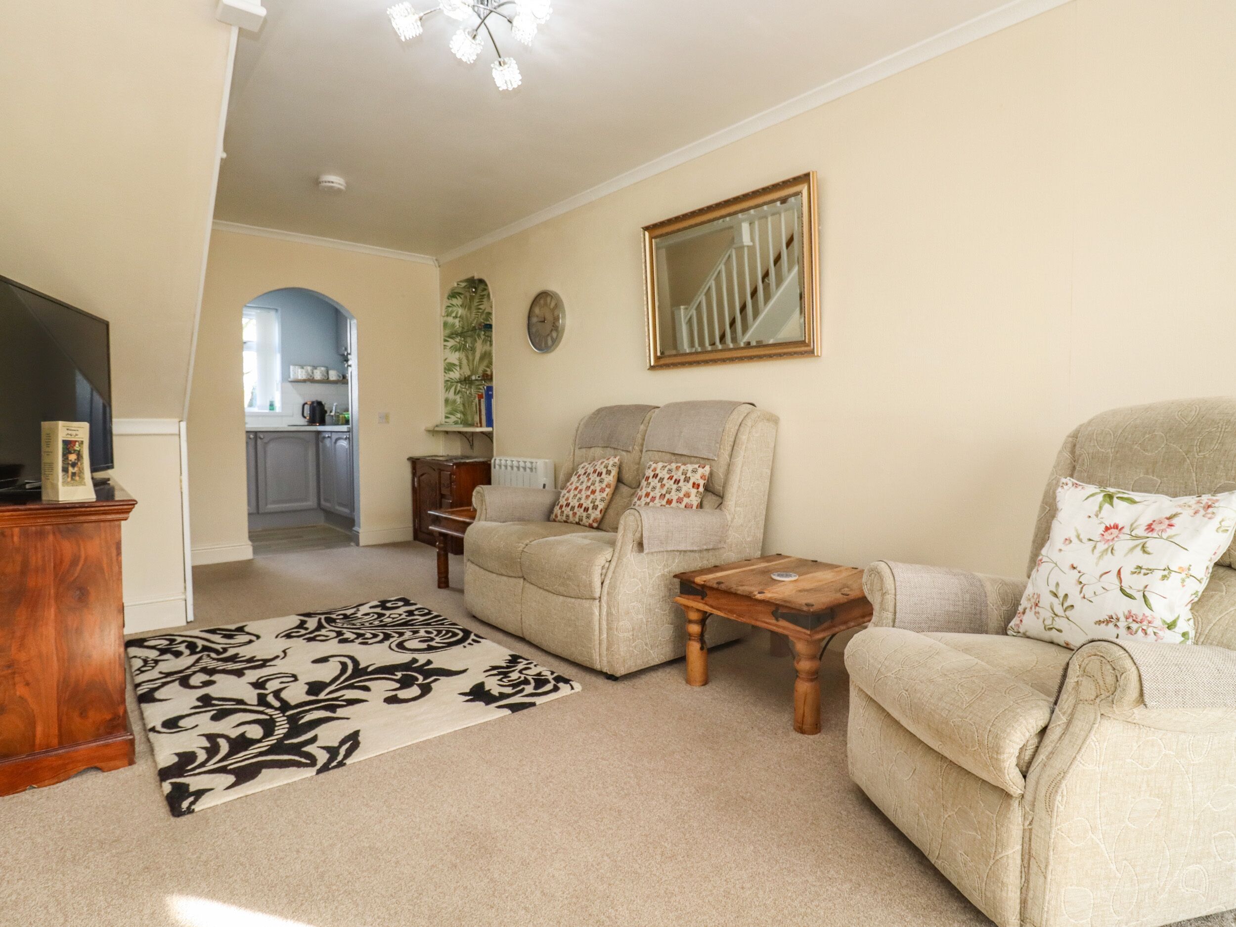 A living room with a sofa, armchair, coffee table, and a television. There is a kitchen in the background.