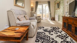 Un salon comprenant un canapé, des fauteuils, une table basse en bois, un tapis à motifs, un téléviseur et un balcon avec des rideaux.