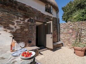 Un bâtiment en pierre avec un toit de chaume, une porte ouverte sur une pièce, une table avec une assiette de fraises et une bouteille de vin.