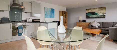 A modern kitchen with white cabinets, a stainless steel range hood, and a glass dining table with four chairs.