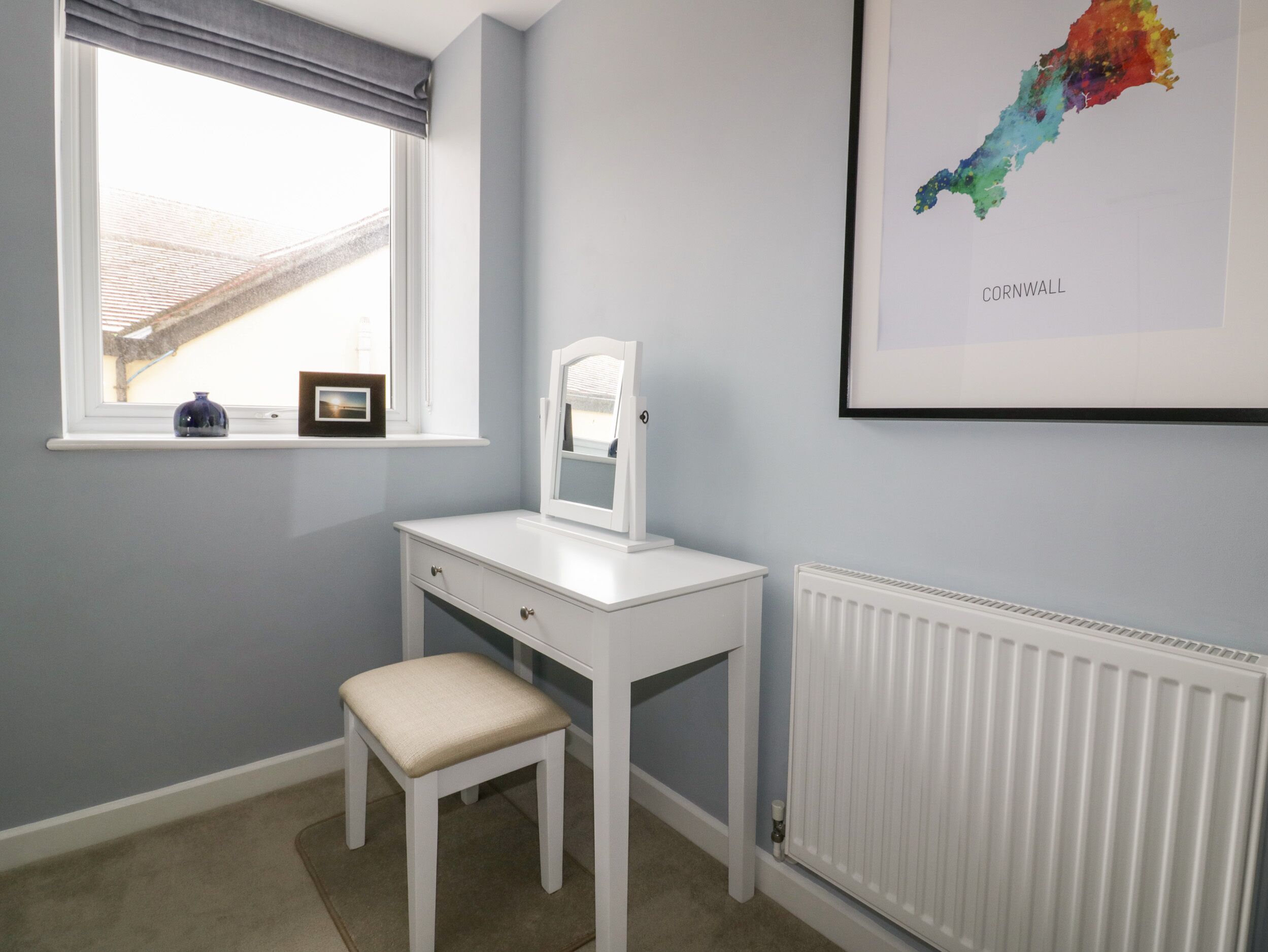 A child's room with a white desk, a chair, a window with a view, a radiator, and a framed map of Cornwall on the wall.