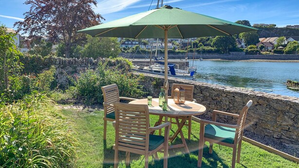 An outdoor patio with a table and chairs, a green umbrella, and a view of a body of water with boats and houses in the background. - Clover Cottage (Plymouth)