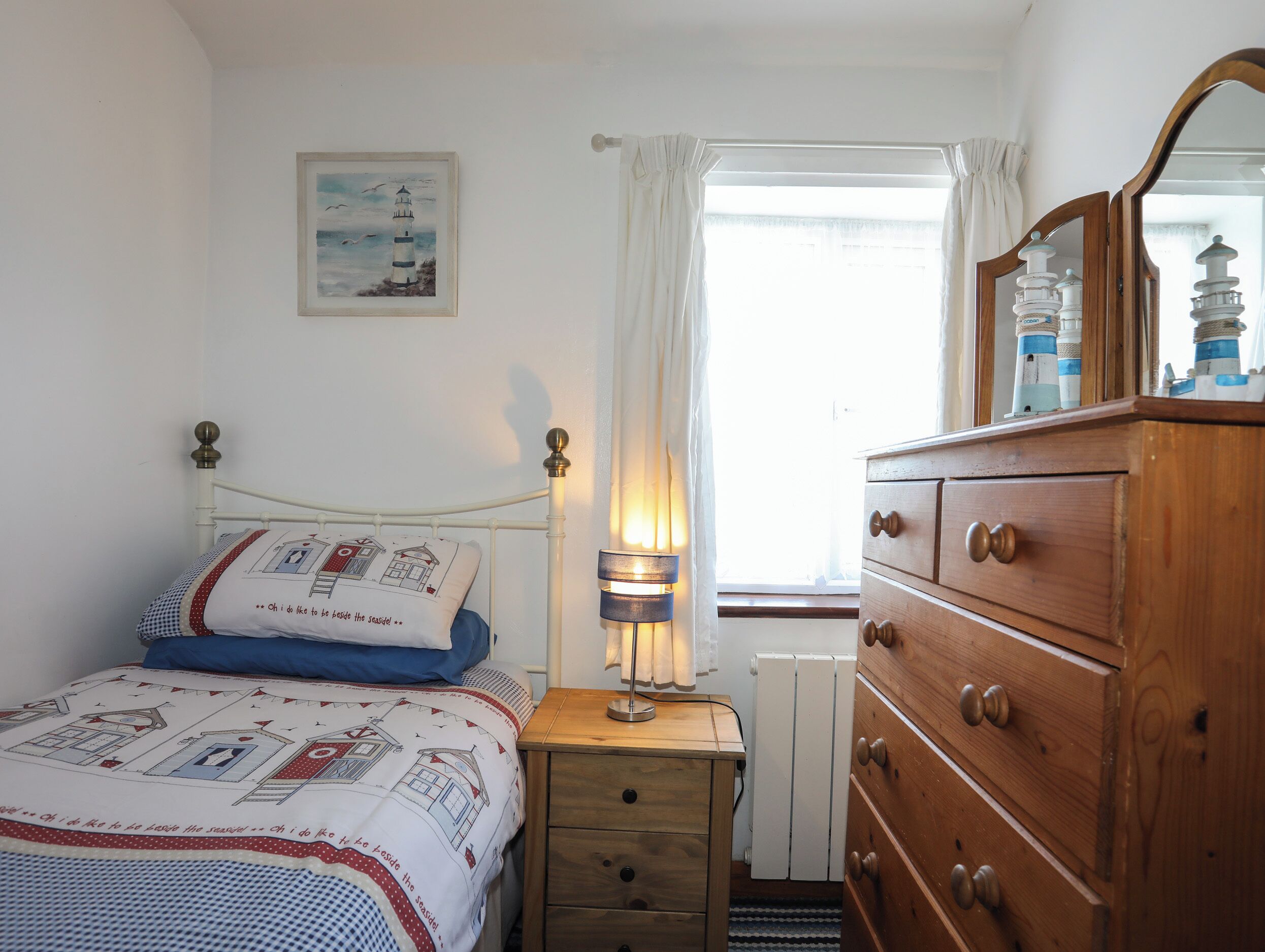 A bedroom with a bed, a bedside lamp, a dresser, a window with curtains, and a painting on the wall.