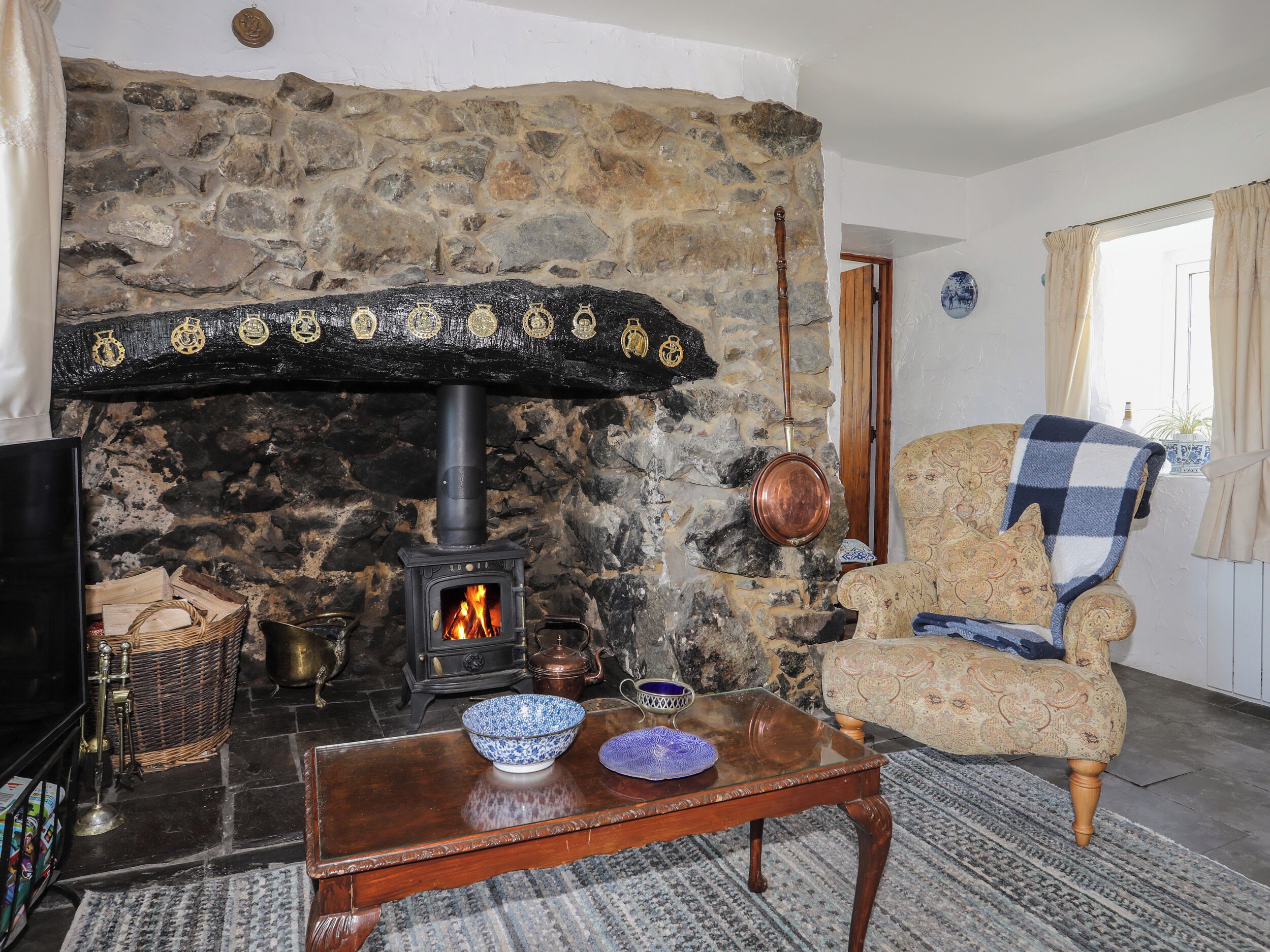 A stone fireplace with a black stove, a wooden table with a bowl, and a patterned armchair.