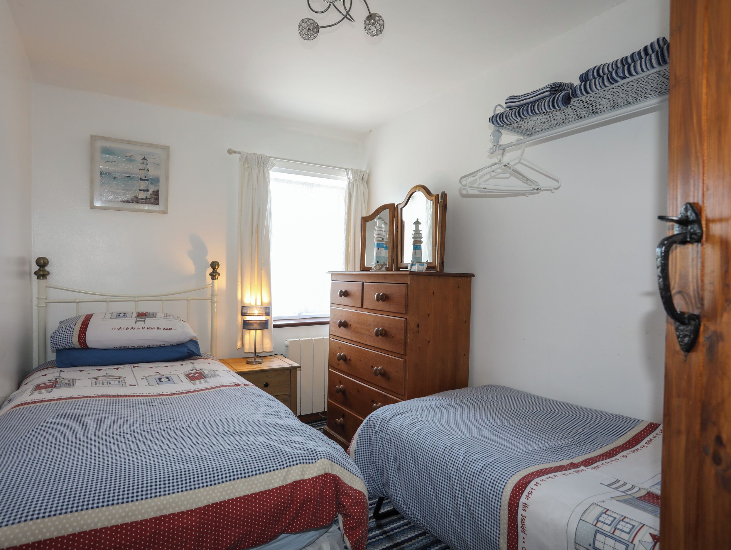 A bedroom with two beds, a wooden dresser, a nightstand, a lamp, a window with curtains, a mirror, and a hanging rack.