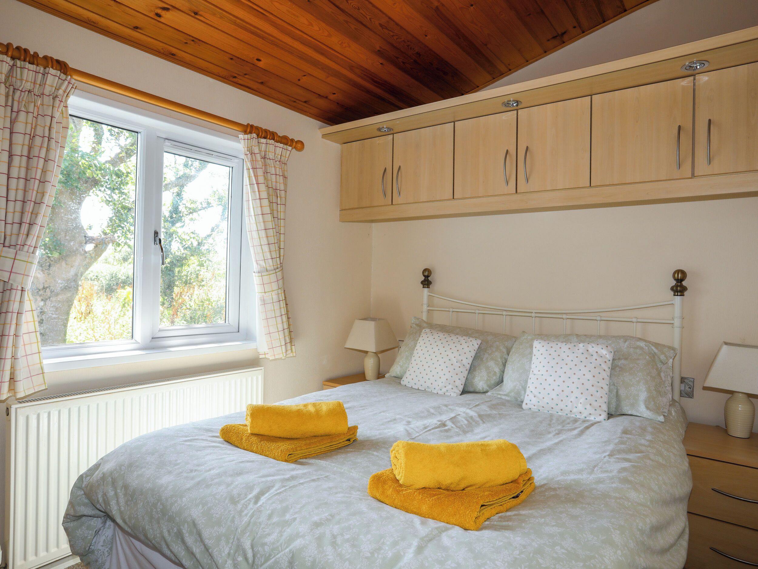 A bedroom with a bed, two bedside lamps, a window with curtains, and a wooden ceiling.