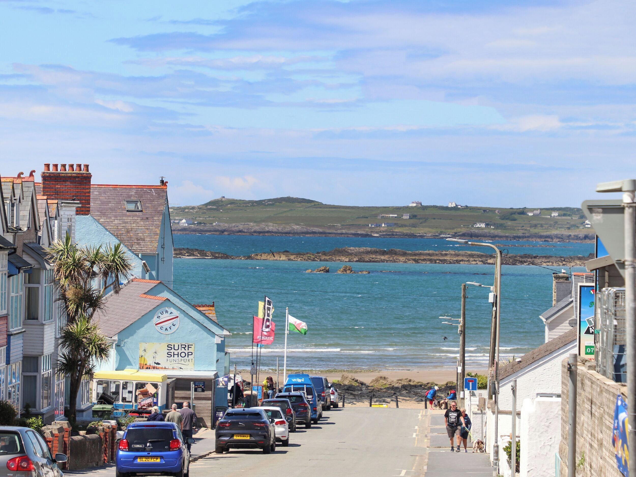 A coastal street with parked cars, a surf shop, and a view of the sea.