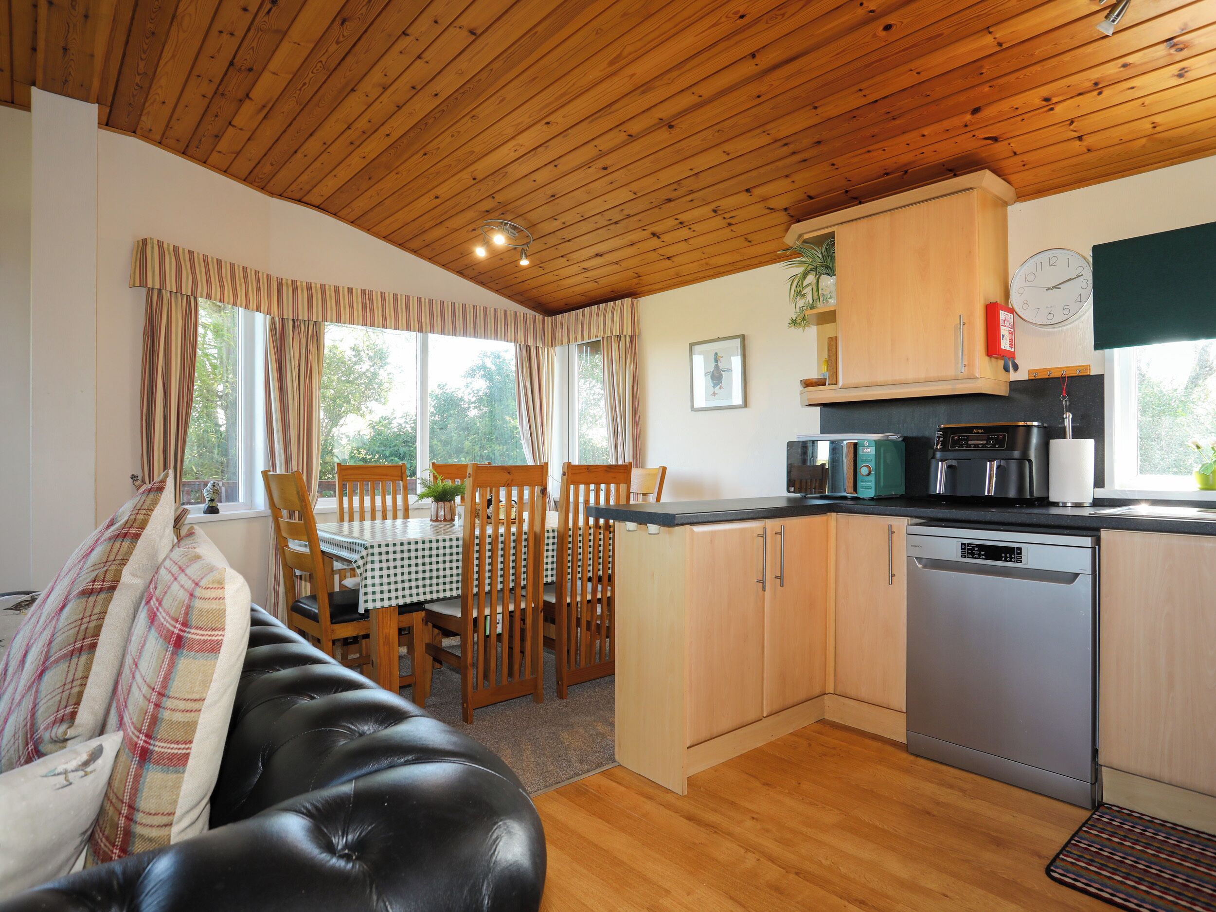 A cozy room with a black leather sofa, wooden dining table, and chairs. A kitchen area with a microwave, toaster, and sink is visible.