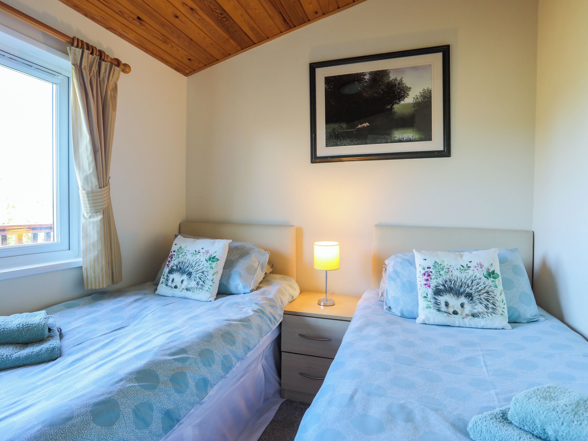 A bedroom with two beds, a window with curtains, a bedside lamp, and a framed picture on the wall.