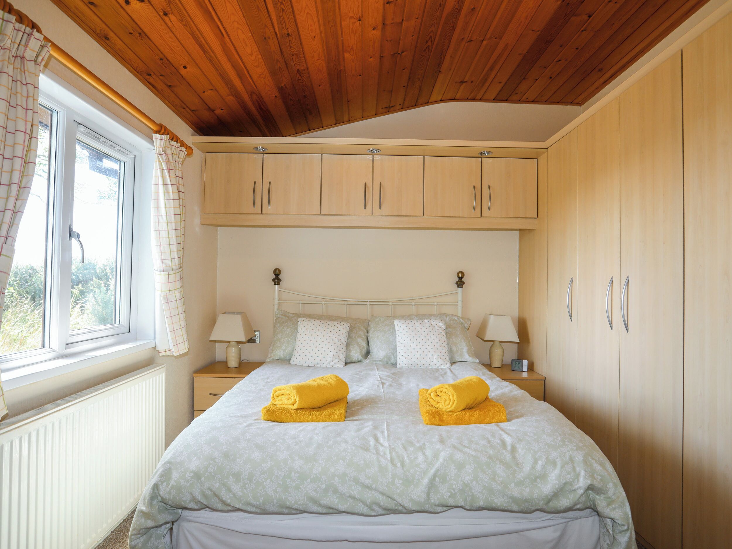 A bedroom with a wooden ceiling, a bed with a floral bedspread, two nightstands with lamps, and a window with curtains.