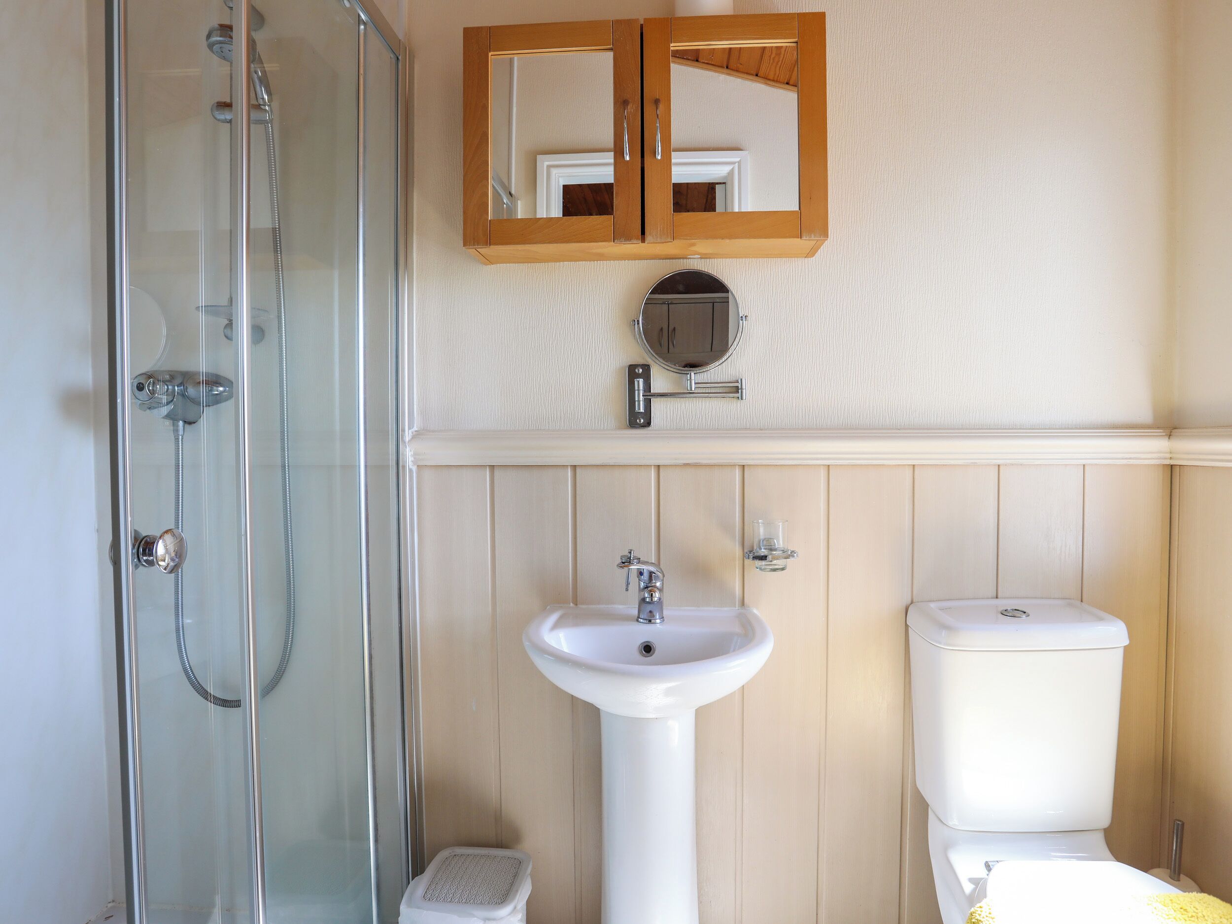 Et badeværelse med en glasvæg omkring brusebadet, et håndvaskarrangement, et toilet og en trækommode med et spejl.
