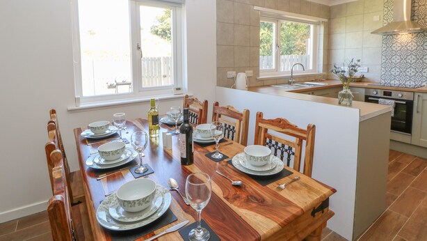 A modern kitchen with a dining area, featuring a wooden table set for a meal, a built-in oven, and a large window providing natural light. - Bankwell Cottage (Newcastle-upon-Tyne)