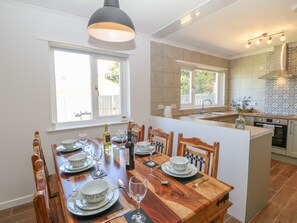 A modern kitchen with a dining area, featuring a wooden table set for a meal, a built-in oven, and a large window providing natural light. - Bankwell Cottage (Newcastle-upon-Tyne)