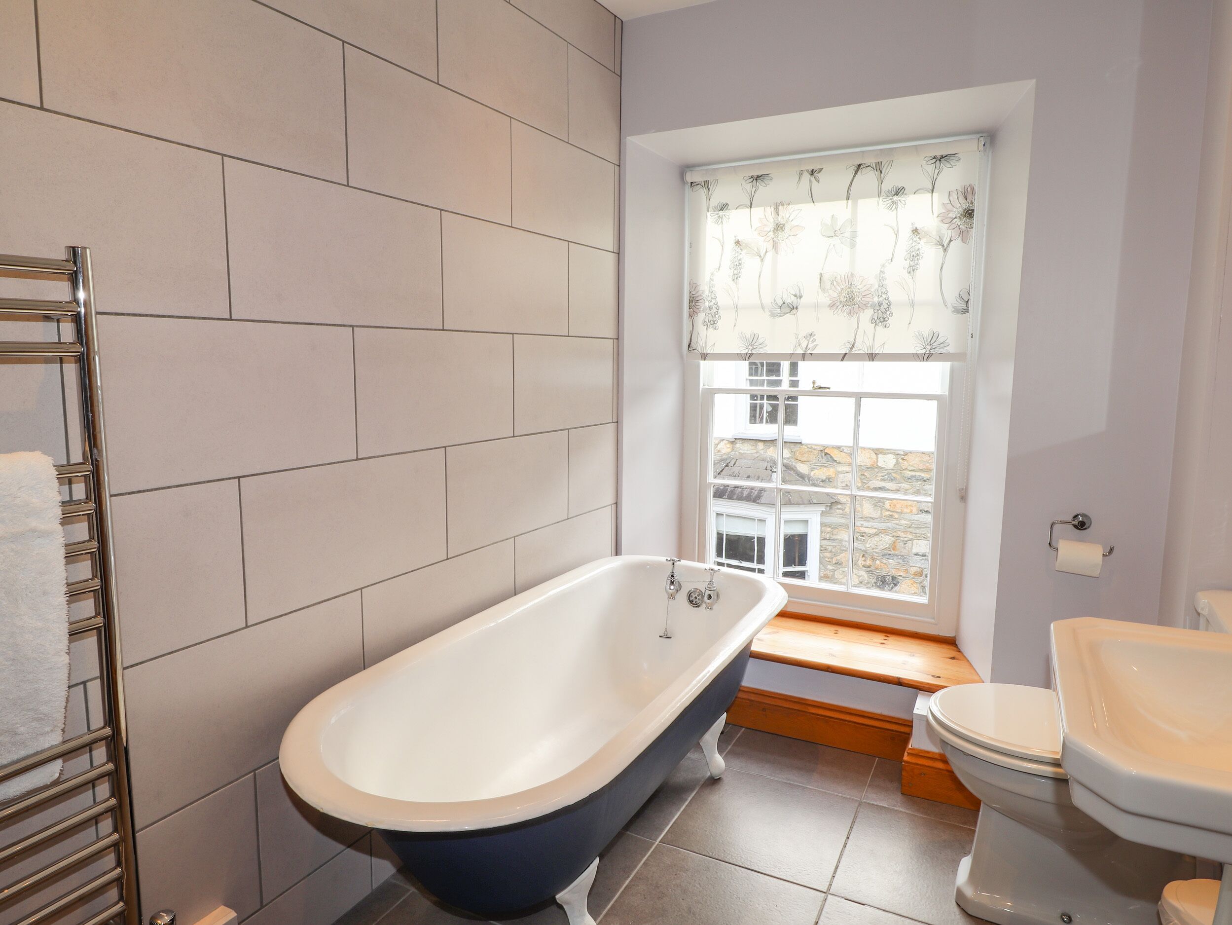 A bathroom with a freestanding bathtub, a toilet, and a window with floral curtains.