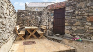 Un patio con paredes de piedra, una mesa de madera y bancos, una puerta de piedra y un pequeño escalón que lleva a un nivel superior.