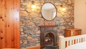 Un dormitorio con pared de piedra, un armario de madera, una chimenea con piñas y un espejo redondo.