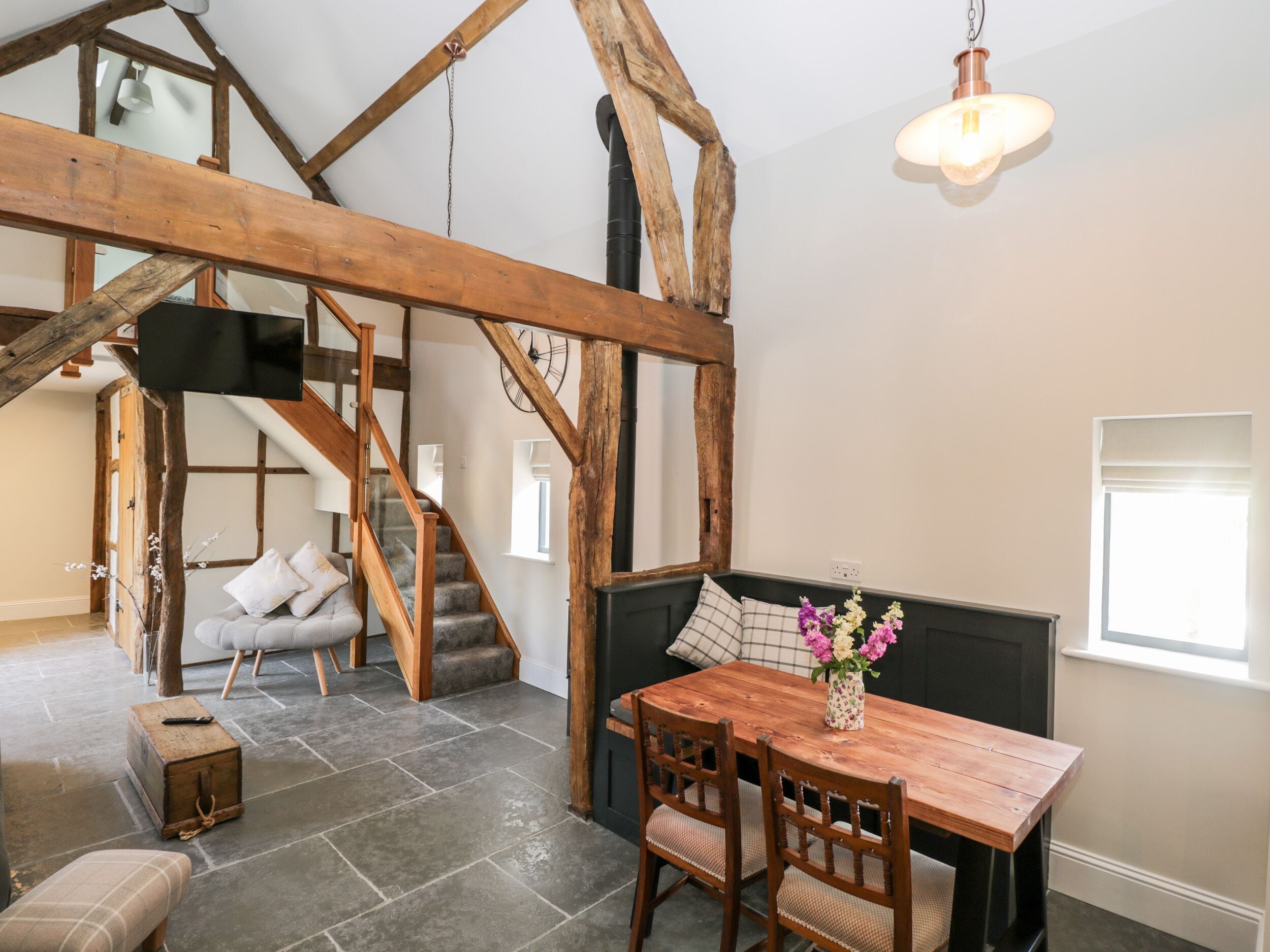 A rustic interior with exposed wooden beams, a stone floor, a wooden dining table with chairs, a bench, and a fireplace.