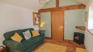 A cozy living room with a green sofa, a yellow rug, a wooden bookshelf, a lamp, and a wood-burning stove. - Yeoman's Cottage (Pickering)