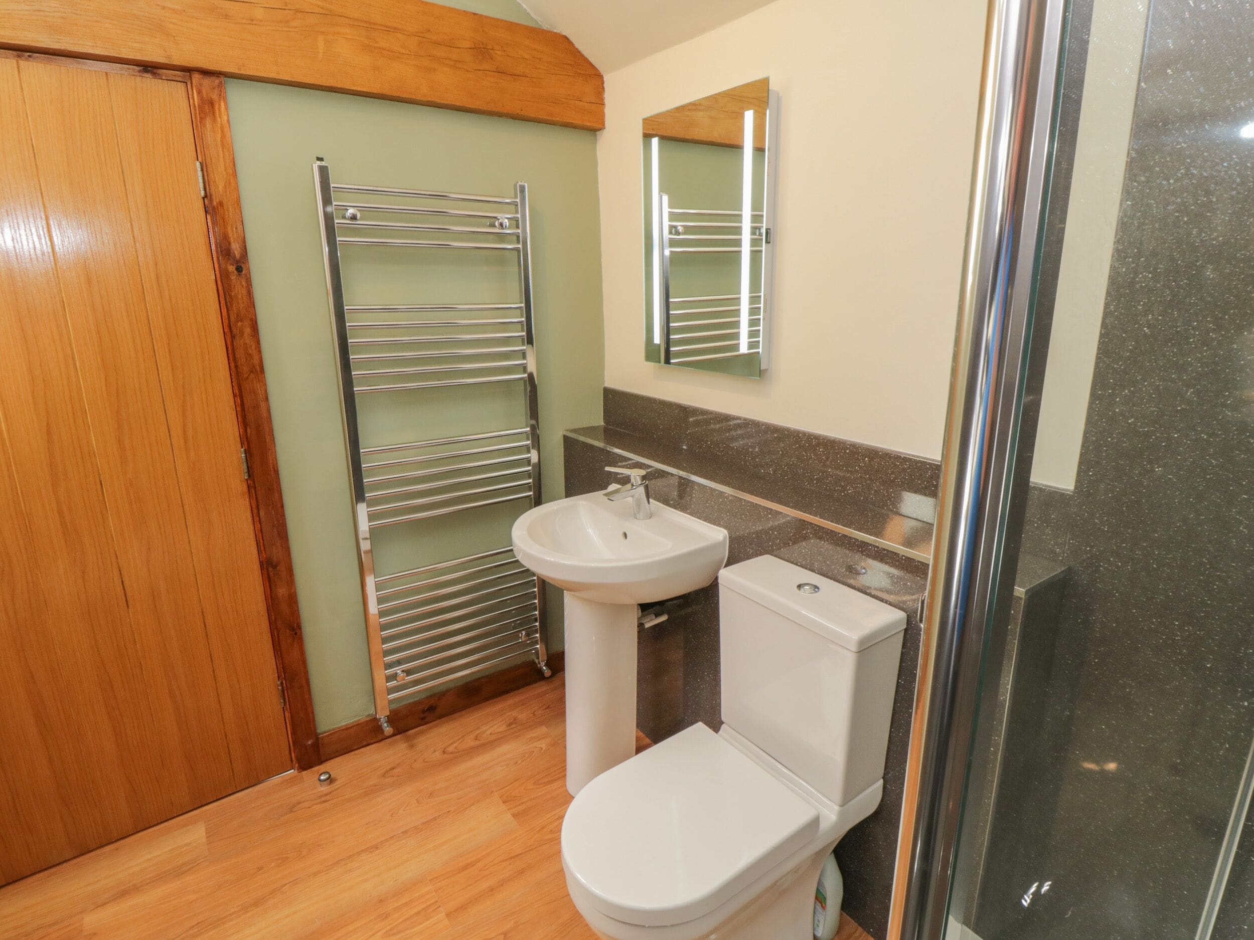 Un baño con piso de madera, un inodoro blanco, un lavamanos, un perchero para toallas y una cabina de ducha de vidrio.