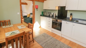 A compact kitchen with white cabinets, a built-in oven, and a microwave. There is a dining table with chairs and a rug on the floor. - Yeoman's Cottage (Pickering)