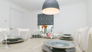 A dining area with a long table set for four, featuring a modern chandelier, white chairs, and a patterned window valance. - Fistral Bay Cottage (Newquay)