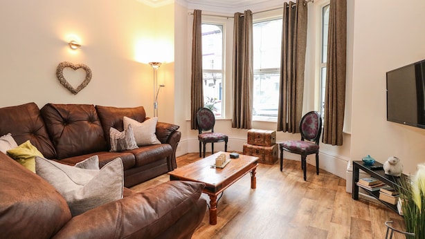 Un salon avec un canapé en cuir marron, une table basse en bois et deux fauteuils. On y trouve une décoration murale en forme de cœur, un lampadaire et un téléviseur fixé au mur.
