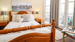 Un lit bien fait, avec une tête de lit en bois, deux tables de chevet avec des lampes, un tableau représentant un paysage portuaire et une vue sur un balcon à travers une grande fenêtre.