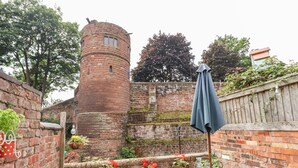 A courtyard with a brick wall, a round tower, potted plants, a table with a tablecloth, and a metal chair. - Tower View Cottage (Chester)