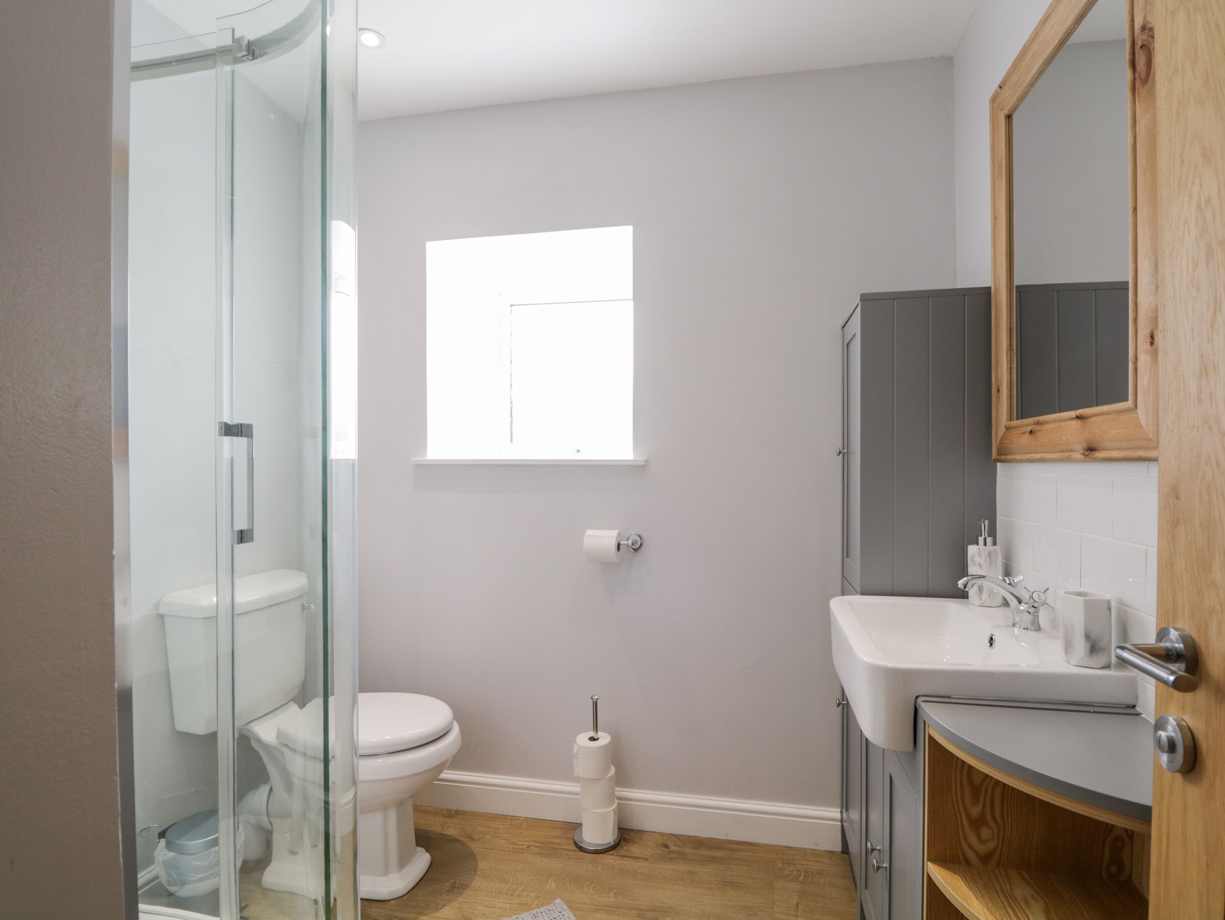 Een moderne badkamer met een douchecabine van glas, een toilet, een wastafel met houten kastje en een raam voor natuurlijk licht.