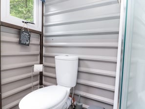 Ett litet, modernt badrum med en vit toalett, en toalettpappershållare och ett fönster med utsikt över grönområden utanför.