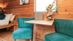 A cozy cabin room with wooden walls, a bed with white bedding, teal upholstered chairs, a small table, and a window with a view. - Shepherd's Hut (Scarborough)