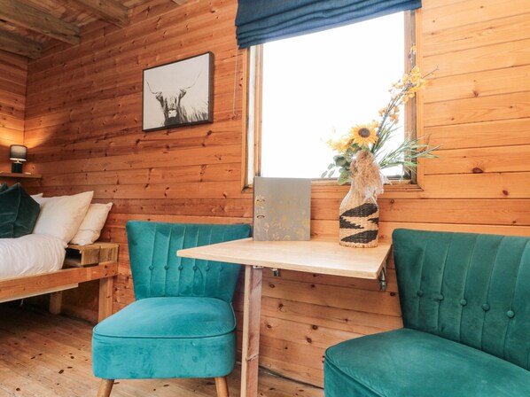 A cozy cabin room with wooden walls, a bed with white bedding, teal upholstered chairs, a small table, and a window with a view. - Shepherd's Hut (Scarborough)