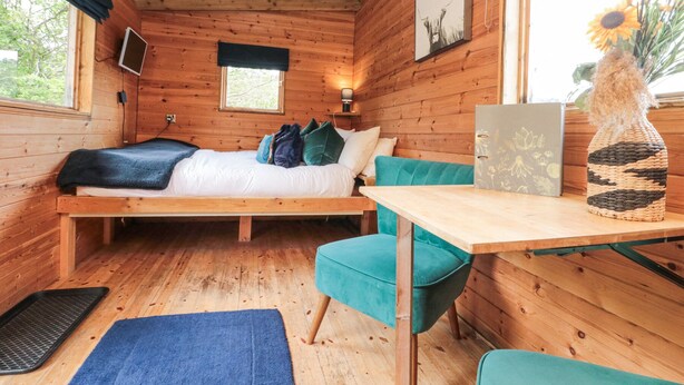 A cozy cabin room with a bed, a small table, a chair, and a window with a view of trees. - Shepherd's Hut (Scarborough)